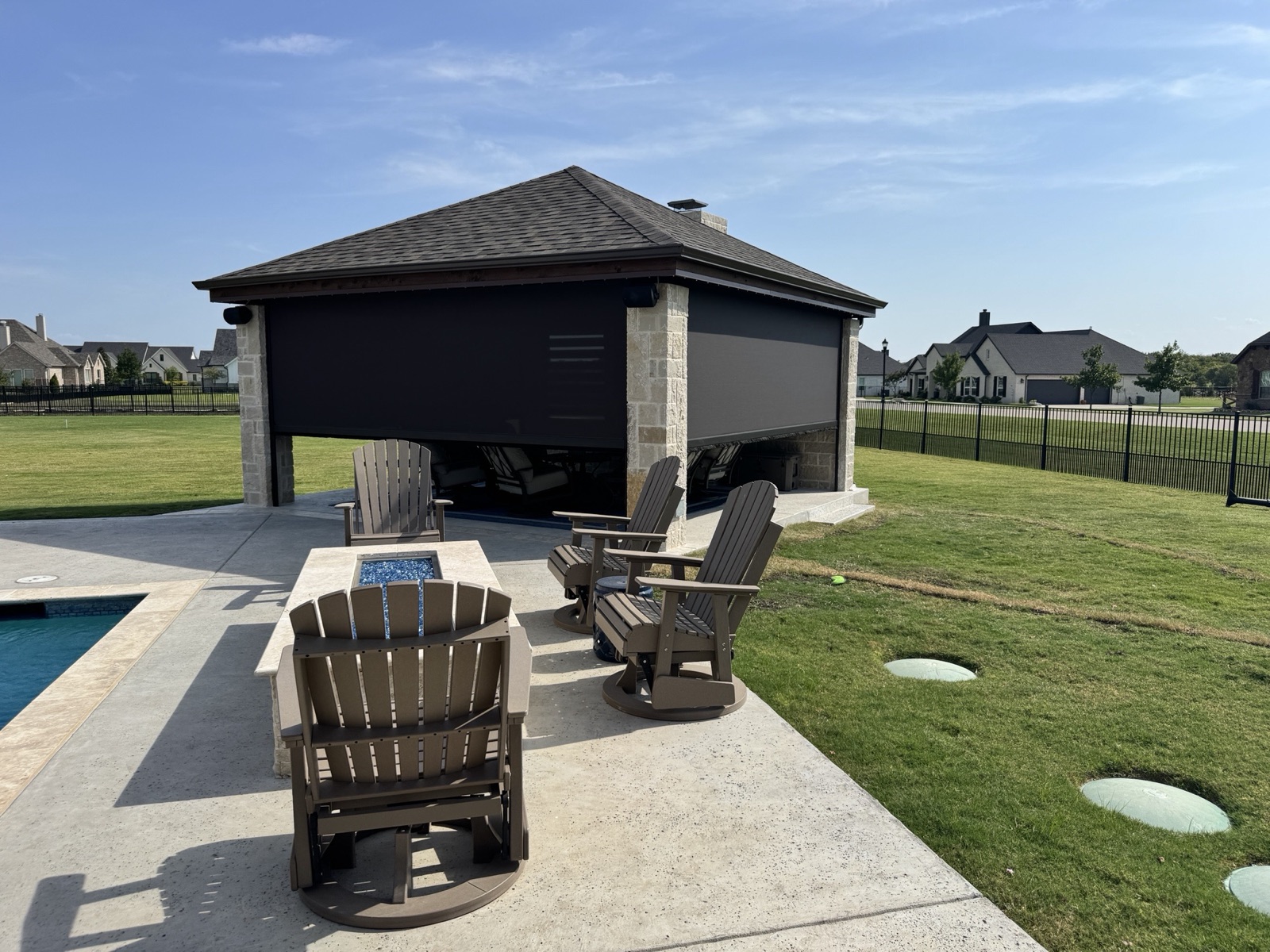 Poolside cabana with dark screens lowered on every side, brown lounge chairs and a fire table in front